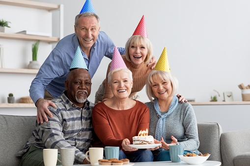 International group of elderly people happy men and women wearing colorful paper hats having birthday party at home, smiling caucasian senior lady holding birthday cake with lit candles, copy space
