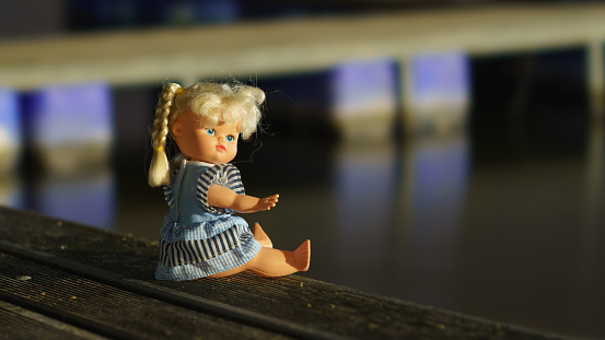 Doll is dressed of blue dress. She has two pigtails. She sits on the wooden bench near the lake. Beautiful girl posing, show direction to the lake by hand. Welcome into deep dive in nature.