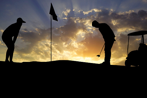 Golfers' hit golf ball toward the hole at sunset silhouetted. Golden morning sky in winter, misty high mountain background.