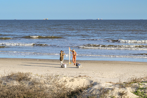 Galveston, Texas, USA - February 2023: Person posing in a swimsuit alongside a surfboard at the water's edge having their picture taken