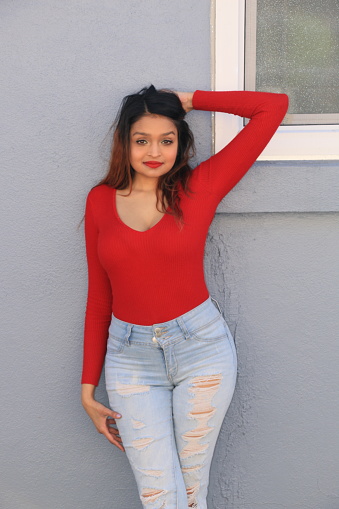 A Fijian model standing in front of a gray stucco wall next to a window. She is wearing long, brown straight hair, a red sweater, makeup, and torn blue jeans.