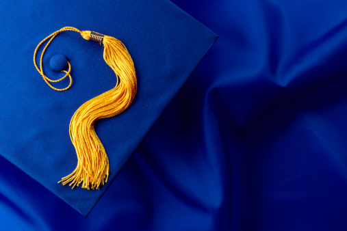 Blue mortarboard and yellow tassel shot on blue graduation gown, space for copy