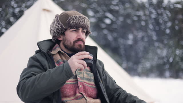 A winter camper wearing a ushanka or hunting hat, winter mountain camper drinking hot coffee by the tent, cold camper drinking hot drink, human in nature away from everything, living free in nature