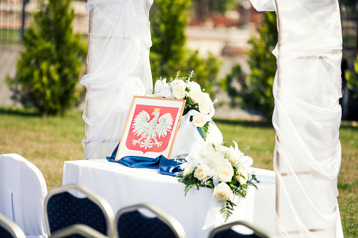 Close-up of an outdoor civil wedding setting - official table, wedding arch, Polish emblem and flowers on a table.