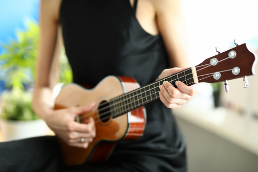 Close-up of person playing melody on ukulele. Female elegant hand pressing on string. Musical talent. Simple black dress on artist. Musical instrument and hobby concept