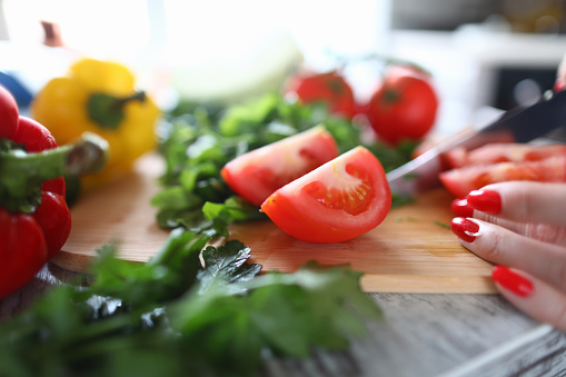 Close-up of person cutting ripe tomato on slices. Red and yellow peppers with greenery on table. Fresh summer vegetables. Healthy eating and tasty lunch concept