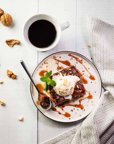 Brownies with ice cream and caramel sauce with a cup of espresso coffee on a white wooden background. Delicious chocolate-nut dessert for breakfast. Top view.