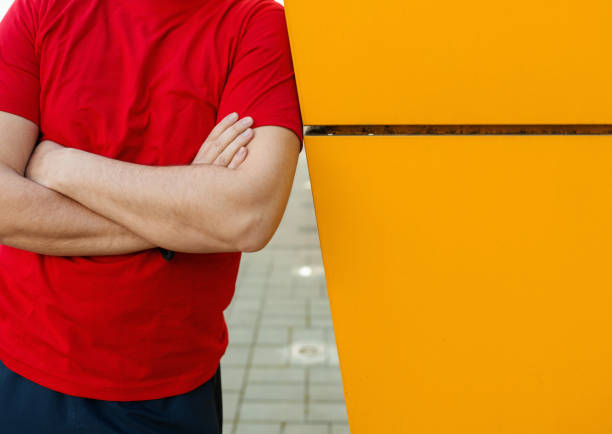 Closeup front view of unrecognizable man leaning against yellow wall while wearing bright red t-shirt.