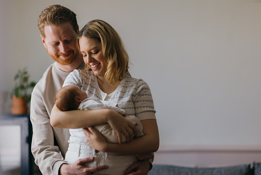 A smiling blonde Caucasian woman holding a baby while her ginger husband is looking at their cute baby and hugging her wife. They are enjoying being parents. (family concept)