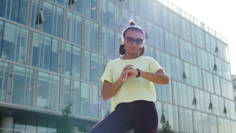 Low angle view of African-American woman in sportswear using headphones and smartwatch on street in morning