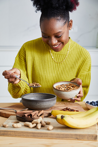 Young adult girl making oatmeal for breakfast, with fresh organic fruit, nuts and grains, at a wooden table in a modern white kitchen, representing a healthy lifestyle, an image with a copy space