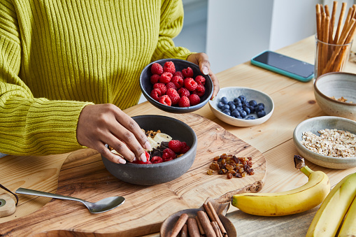 Young adult girl making oatmeal for breakfast, with fresh organic fruit, nuts and grains, at a wooden table in a modern white kitchen, representing a healthy lifestyle, an image with a copy space