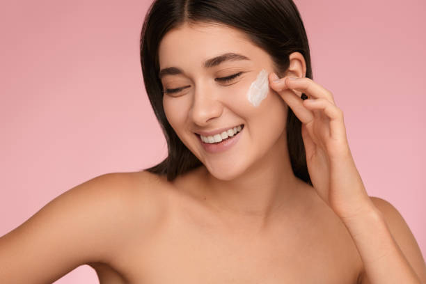 istock Smiling young lady applying moisturizing cream on face in pink studio 1479201567