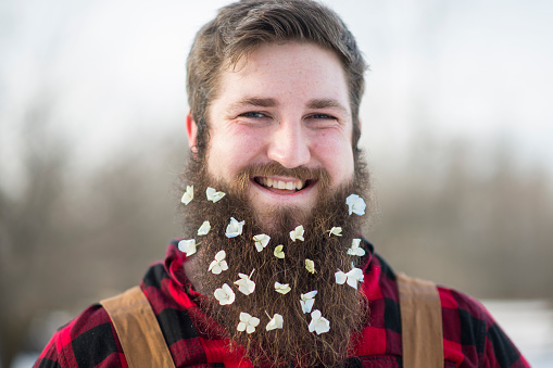 Portrait of a smiling man with delicate white flowers decorating his long brown beard. He is wearing black and red plaid shirt and work coveralls.