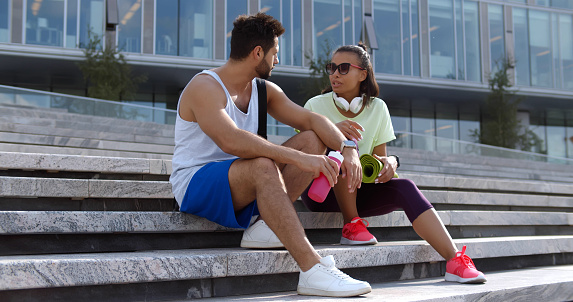 Fit diverse couple taking rest after workout outdoors, sitting on urban stairs drinking water and chatting. African-American woman and indian man relax on staircase outdoors after workout