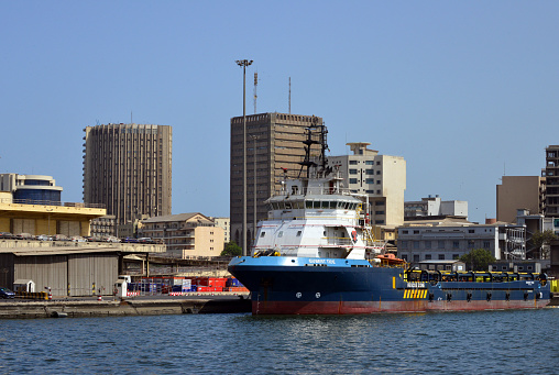 Dakar, Senegal: Gubert Tide (IMO: 9543823), platform supply vessel (PSV) / offshore supply ship (OSS), a ship specially designed to supply offshore oil and gas platforms - They ensure the security of the drilling rigs and their supply of material and are used for towing to new locations. They are equipped with powerful machinery to withstand the adverse weather conditions that often prevail in offshore rig areas. Usually these ships have bow thrusters, as shown  by the two 'propeller' markings on the bow of the Gubert Tide - Autonomous Port of Dakar (PAD) with business district towers in the background (BCEAO, Fahd, Azur...)