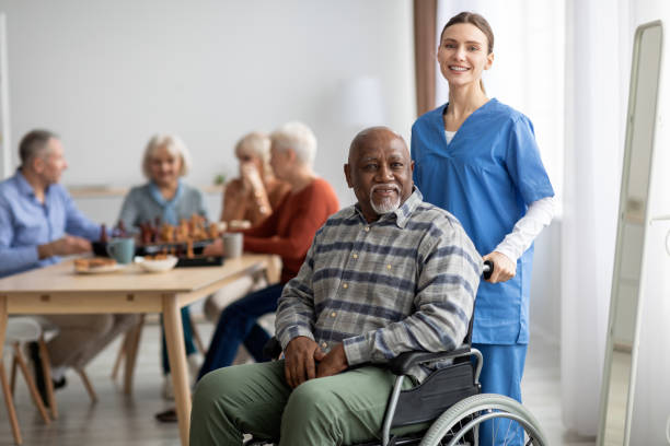 Happy older patient on wheelchair with female nurse Happy black man older patient on wheelchair with female nurse smiling at camera, group of senior people sitting on couch on background, nursing home interior, healthcare for elderly people concept common room stock pictures, royalty-free photos & images