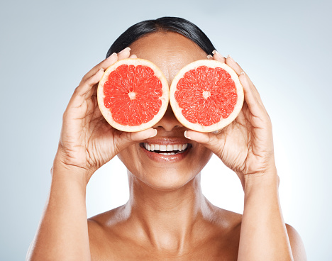 Funny, fruit and woman use grapefruit for a joke covering her eyes isolated against a studio blue background. Health, beauty and skincare model smiling and laughing with juicy citrus