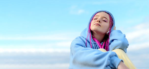 Portrait of a caucasian teenage girl with pink braids using wireless headphones against the blue sky.Technology,summer concept.Generation Z style.Copy space for text.
