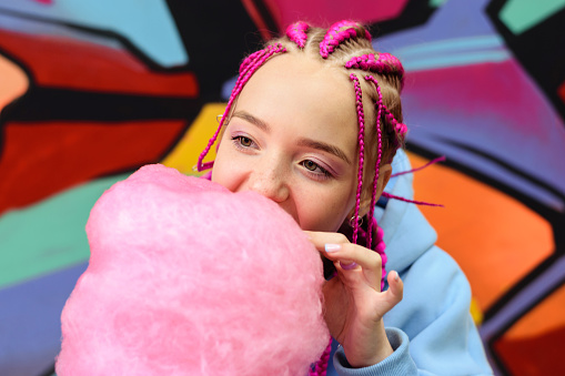 Caucasian teenage hipster girl with pink braids eats cotton candy against the background of a multicolored street wall.Summer concept.Generation Z style.