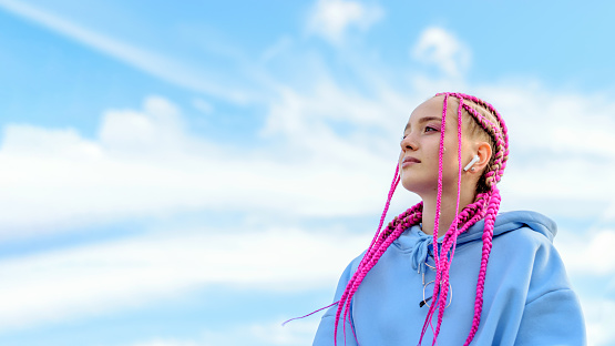 Portrait of a caucasian teenage girl with pink braids using wireless headphones against the blue sky.Technology,summer concept.Generation Z style.Copy space for text.