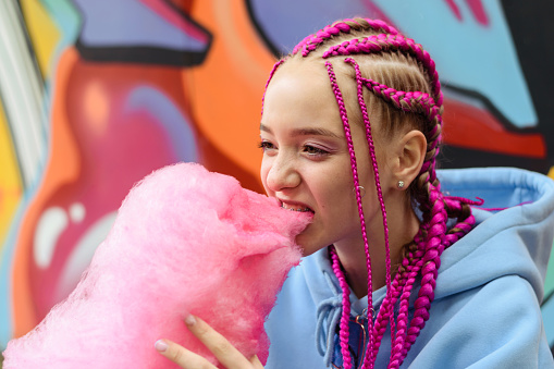Caucasian teenage hipster girl with pink braids eats cotton candy against the background of a multicolored street wall.Summer concept.Generation Z style.