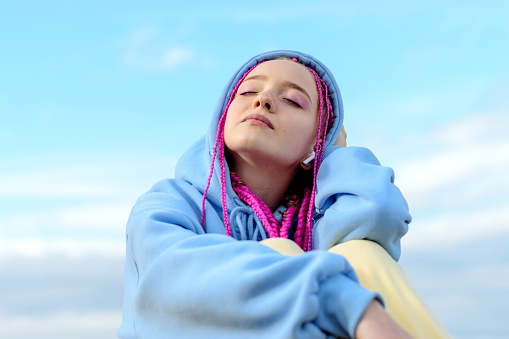 Portrait of a caucasian teenage girl with pink braids using wireless headphones against the blue sky.Technology,summer concept.Generation Z style.