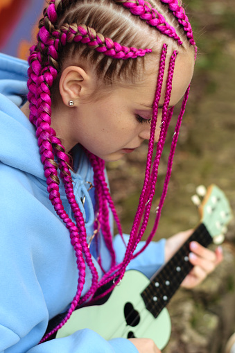 Caucasian teenage hipster girl with pink braids plays the ukulele on the street.Summer concept.Generation Z style.