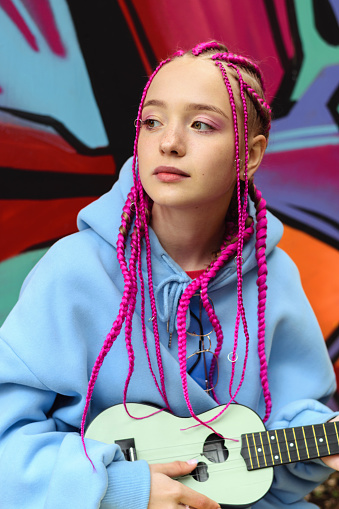 Caucasian teenage hipster girl with pink braids plays the ukulele against the background of a multicolored street wall.Summer concept.Generation Z style.