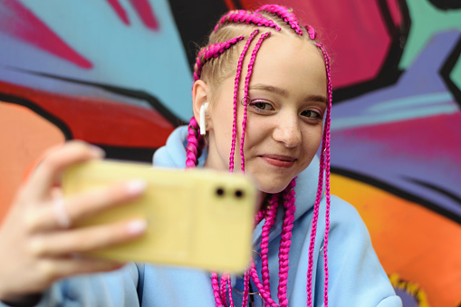 Caucasian teenage hipster girl with pink braids using a smartphone against the background of a multicolored street wall.Summer concept.Generation Z style.Social media concept.