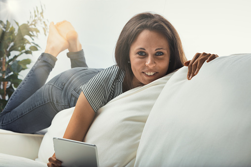 Young woman lounging on the edge of the backrest of a white couch, lazily contorted because of her fit body, looks at something on the invisible screen of a digital tablet - maybe a video, a book or a