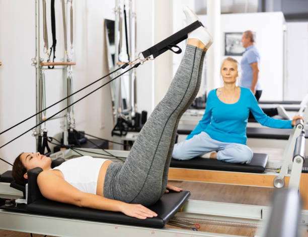 istock Young European female training lower back while laying and pulling with legs bands of Pilates reformer machine bed at gym. Pilates and healthy lifestyle concept 1479283941