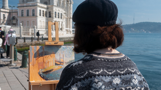 The young female painter artist look at the Ortakoy Mosque on the shore of Ortaköy and paints it front of her easel. Besiktas, Istanbul, Turkey