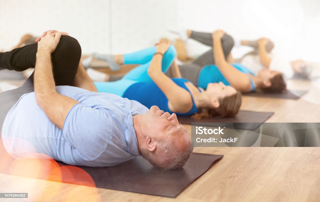 Elderly man doing glutes stretching exercises at training in gym Concentrated elderly man doing stretching exercises during group training in gym, lying on mat and pulling knee toward shoulder to relax glutes and release tension from sciatic nerve Gym Stock Photo