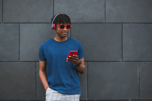 Young adult with red sunglasses and headphones listening to music with smartphone, standing on a wall with copy-space.