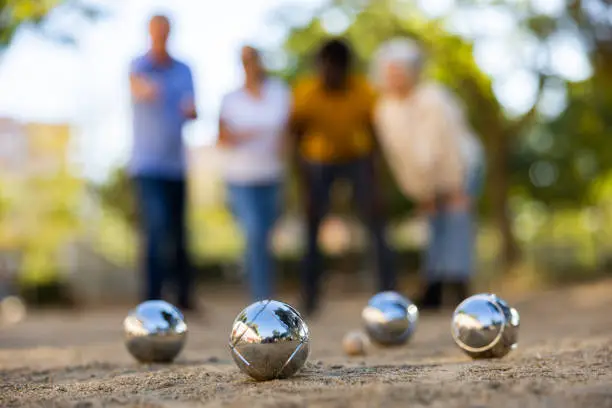 View of petanque shiny silver metal balls laying on the ground outside in the park with group of people standing behind on a sunny day