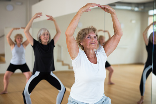 Mature women who are engaged in the dance section in the studio at a group lesson perform a plie squat, being in the position 
of a ballet stand