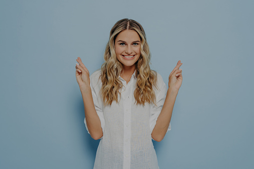 Young lovely caucasian blonde female with beaming smile raising hands and crossing fingers with hope and luck gesture, anticipating important news, isolated over blue studio background