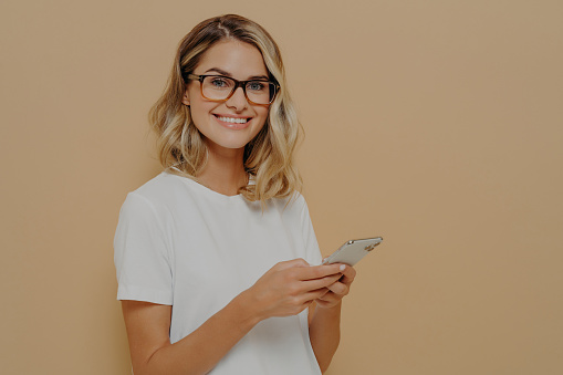 Young blonde smiling businesswoman in casual wear and eyeglasses using using mobile phone, checking emails on smartphone while spending time indoors, looking at camera with positive facial expression