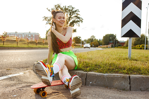 Glad young female hipster sitting on road with skateboard and looking at camera while enjoying summer sunset