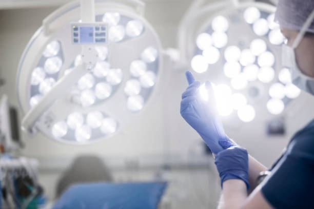 Female surgeon putting on surgical gloves in operation room Over the shoulder view of a female surgeon preparing for operation. Close-up of a woman doctor in surgical gown putting on surgical gloves in operation room. putting on latex gloves stock pictures, royalty-free photos & images