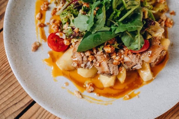 There is a dish in the popular restaurant. We can see a lot of green and orange sauce. There is a meat and tomatoes on the white plate on the wooden table. There are a lot of slices of potato.