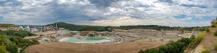 Open pit quarry for mining limestone in the Sint Pietersberg (Mount Saint Peter) in Limburg near Maastricht during an overcast summer day in The Netherlands. On the left the processing plant for creating cement and concrete for the building industry.