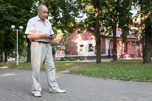An elderly man walks alone in the park in the summer. A modern retired businessman in a white shirt and trousers writes a message and studies information on a mobile phone.