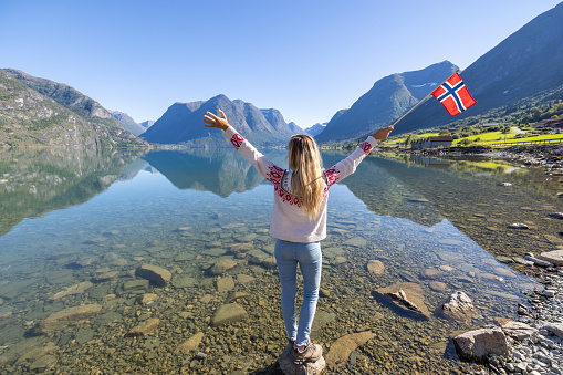 Female holding Norwegian flag against the mountain landscape in the fjords. Beautiful sunny day, reflection on water.