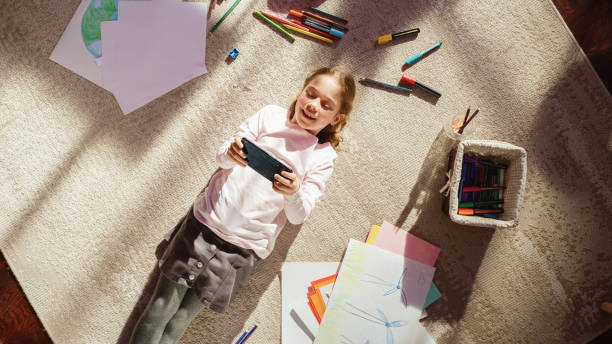istock Top View Home: Happy Little Girl Using Smartphone While Lying on the Floor. Joyful Sunny Day and Clever Smiling Child Playing Smart Video Games, Using e-Learning Apps on Mobile Phone. 1479315744