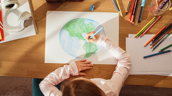 Top View: Little Girl Drawing Our Beautiful Planet Earth. Very Talented Child Having Fun at Home, Imagining Our Home Planet as a Happy Place with Clean, Sustainable Living. Cozy Sunny Day.