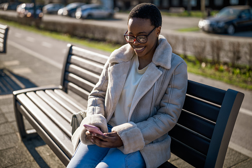 Outdoor portrait of happy african-american woman on sunny day. She is sitting on bench in the street and messaging on smartphone.
