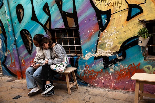 Athens, Greece – October 23, 2022: The young girls sitting on a bench in front of a graffiti street art in Athens, Greece.