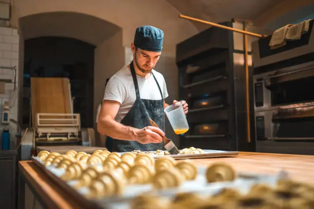 Caucasian baker applying egg wash on to raw pastries on a baking trays before going into an oven in a small artisan bakery.